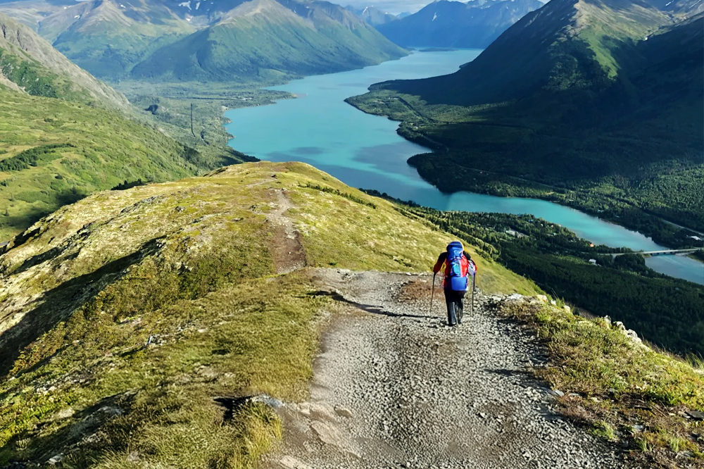 A hiker with a red backpack walking along a ridge trail overlooking turquoise rivers and rugged mountains under a clear blue sky in Cooper Landing, Alaska.